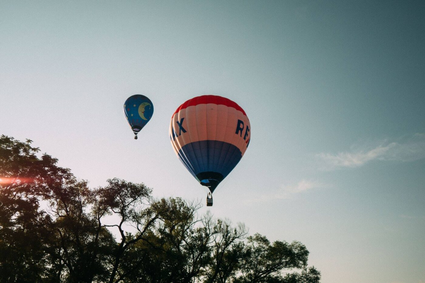 Two hot air balloons floating in the sky. The smaller balloon is blue with a yellow moon and stars. The larger balloon features the Remax logo